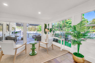 Bright coastal-style sunroom with two white armchairs, green ceramic side table, potted palm and large windows opening to a tropical backyard patio with wicker seating.