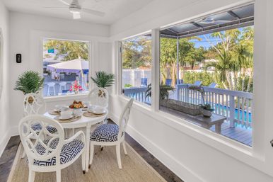 Bright coastal sunroom breakfast nook with a white round table set for coffee and pastries, four blue-patterned chairs, potted palms, and large windows overlooking a deck, swimming pool and tropical backyard with palm trees and umbrella.