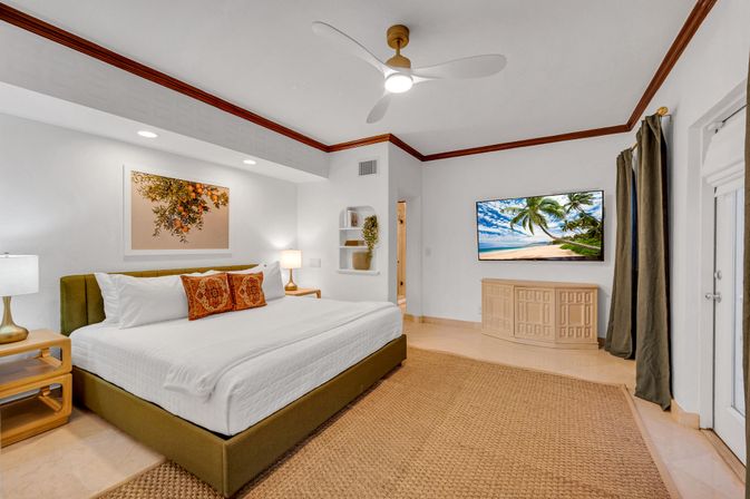 Bright coastal resort suite bedroom with king bed in white linens and patterned throw pillows, ceiling fan, bedside lamps and tables, woven area rug, wall-mounted TV showing a palm-lined beach, wood-trimmed crown molding and sliding glass door with curtains.