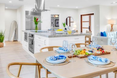 Bright open-concept coastal-style kitchen with white cabinets and marble island, stainless refrigerator, wooden dining table set with blue-and-white napkins, wine glasses and a colorful charcuterie board