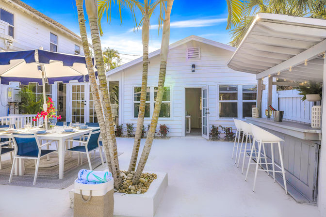 Sunny tropical backyard patio of a white beach cottage featuring palm trees, a blue-chaired outdoor dining set with umbrella, and a coastal bar with white stools