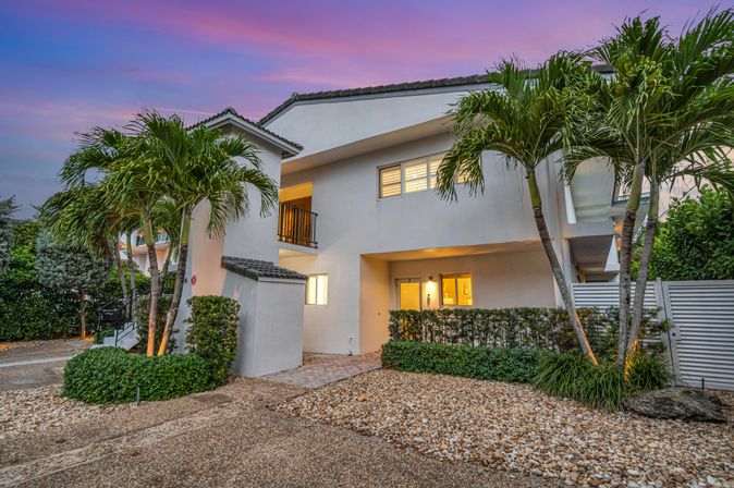 Twilight view of a modern white tropical townhouse with palm trees, pebble landscaping, a small balcony and warm interior lights beneath a pink sunset sky.