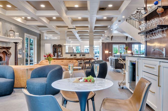 Bright open-concept luxury lounge with coffered ceiling, marble bistro table set with champagne flutes and fruit, blue and leather seating, and a bar with hanging glassware.