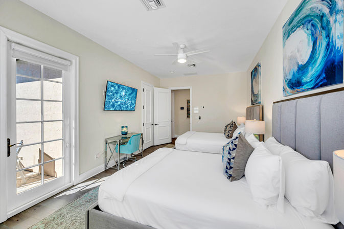 Bright coastal-themed guest room with two queen beds, gray upholstered headboards, crisp white linens, blue wave artwork, wall-mounted TV, glass patio door, small desk with turquoise chair, and ceiling fan.