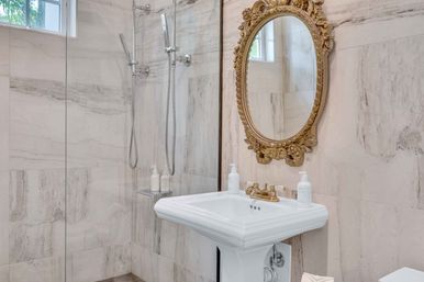 Luxury white marble bathroom with glass walk-in shower, twin handheld showerheads, classic pedestal sink and ornate gold oval mirror.
