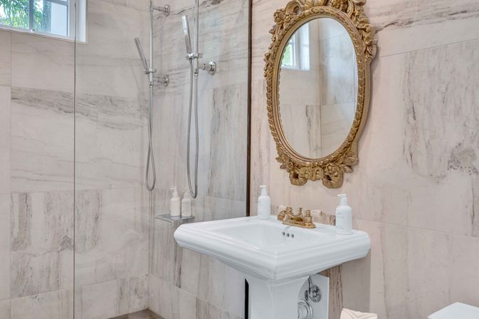 Luxury white marble bathroom with glass walk-in shower, twin handheld showerheads, classic pedestal sink and ornate gold oval mirror.