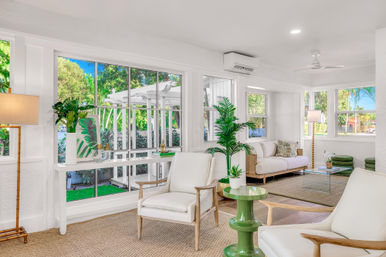 Sunlit coastal-style living room with large windows overlooking lush tropical greenery and a white pergola; airy white walls, mid-century wood-frame armchairs and sofa, green accent table, potted plants, woven jute rugs, glass coffee table, floor lamps, ceiling fan and wall-mounted AC.