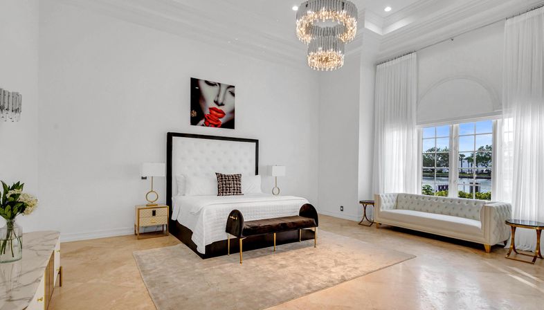 Bright luxury waterfront master bedroom with white tufted king bed, velvet bench at foot, crystal chandelier, tufted sofa by large arched window overlooking water, neutral rug and modern bedside tables