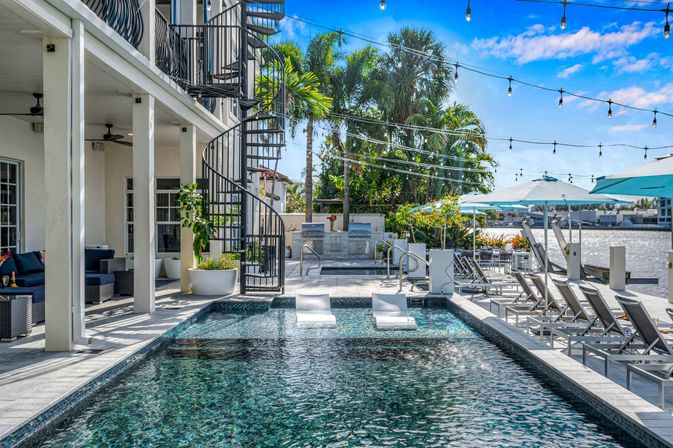 Waterfront resort-style pool with in-pool loungers, black spiral staircase, palm trees, string lights and rows of sunbeds and umbrellas on a sunny day