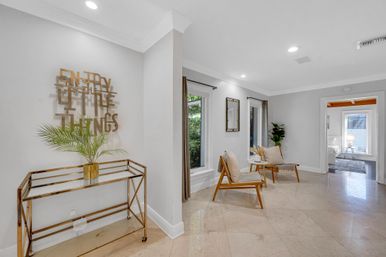 Bright modern home sitting area with polished marble floors, two mid-century wooden accent chairs with cushions and small table, a tall potted plant, and a gold metal console topped with a palm under wall art reading "Enjoy the little things."
