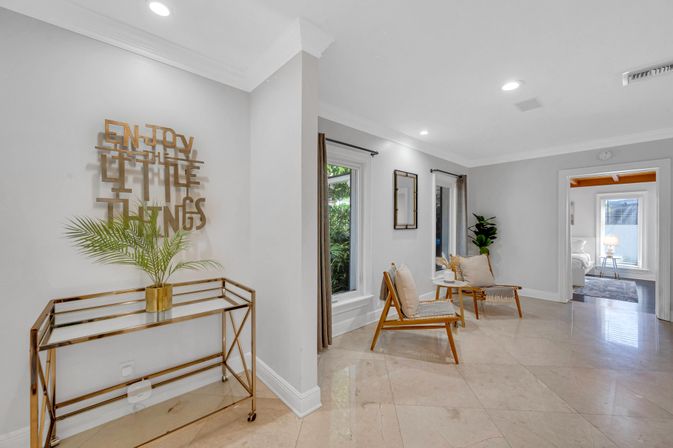 Bright modern home sitting area with polished marble floors, two mid-century wooden accent chairs with cushions and small table, a tall potted plant, and a gold metal console topped with a palm under wall art reading "Enjoy the little things."