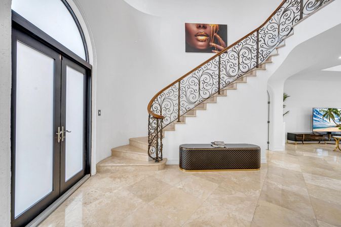 Bright luxury foyer with curved marble staircase and ornate wrought-iron railing, arched frosted-glass double doors, quilted bench and gold-lip wall art.