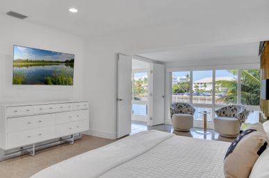 Bright modern waterfront bedroom with white dresser and wall TV, open double doors to a sunlit seating nook with two patterned chairs and large windows overlooking a marina, palm trees, and waterfront homes — coastal vibes.