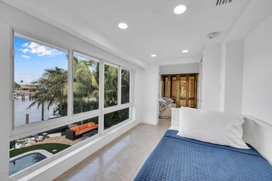 Sunlit coastal bedroom nook with blue quilted bed and large windows overlooking a palm-lined canal, pool and orange outdoor sofa