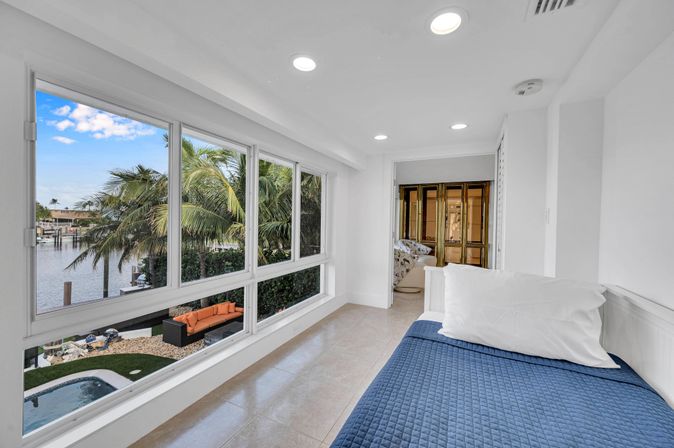 Sunlit coastal bedroom nook with blue quilted bed and large windows overlooking a palm-lined canal, pool and orange outdoor sofa