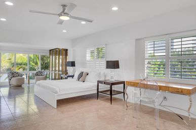 Bright modern coastal master bedroom with white platform bed, dark bedside tables and lamps, clear acrylic desk and chair, seating area by large windows showcasing palm trees, ceiling fan and tiled floor.