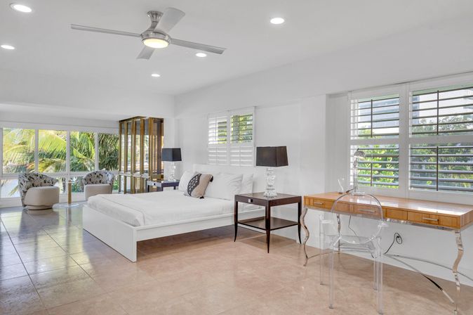 Bright modern coastal master bedroom with white platform bed, dark bedside tables and lamps, clear acrylic desk and chair, seating area by large windows showcasing palm trees, ceiling fan and tiled floor.