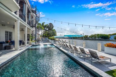 Sunlit waterfront pool beside a modern coastal terrace with lounge chairs, turquoise umbrellas, string lights and a spiral staircase