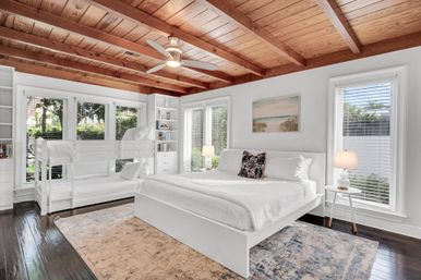 Sunlit coastal-inspired bedroom with exposed wooden beam ceiling, white platform king bed, twin bunk beds, built-in shelves, large windows with blinds, ceiling fan and dark hardwood floors with area rug.