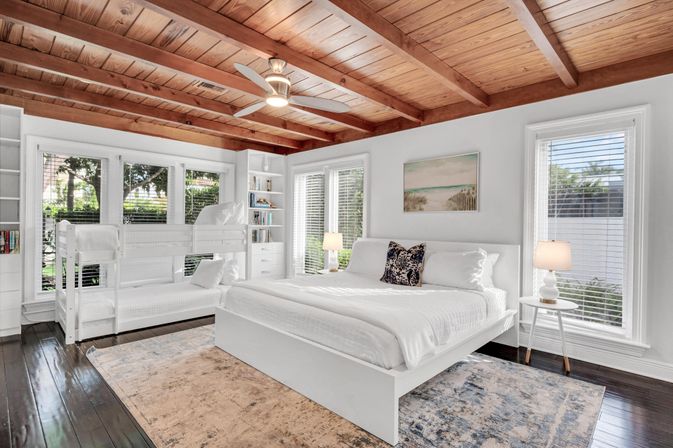 Sunlit coastal-inspired bedroom with exposed wooden beam ceiling, white platform king bed, twin bunk beds, built-in shelves, large windows with blinds, ceiling fan and dark hardwood floors with area rug.