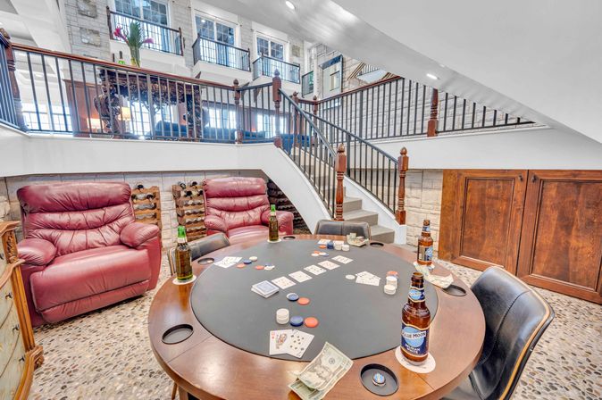 Basement game room with round poker table set with cards, chips and cash, leather recliners, beer bottles, wine racks and a curved staircase leading to an upper loft — cozy entertainment space.