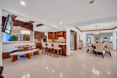 Bright open-concept luxury kitchen and dining area with marble floors, dark wood cabinets, island with three upholstered bar stools, curved breakfast nook with bench seating, stainless steel refrigerator, and gold orb chandelier over the dining table.