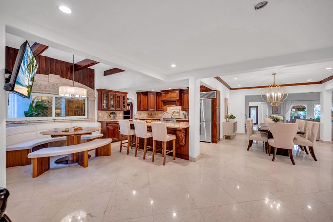 Bright open-concept luxury kitchen and dining area with marble floors, dark wood cabinets, island with three upholstered bar stools, curved breakfast nook with bench seating, stainless steel refrigerator, and gold orb chandelier over the dining table.
