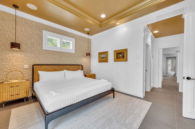 Bright modern coastal bedroom with king bed, woven textured accent wall, crisp white linens, wood nightstands, hanging pendant lights, plantation shutters with palm-leaf view and gold-toned tray ceiling