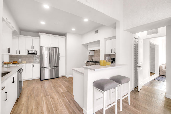 Bright modern white kitchen with stainless steel French-door refrigerator, breakfast bar with two gray stools, marble-look countertops, neutral tile backsplash, wood-look tile floor and recessed lighting in an open layout.