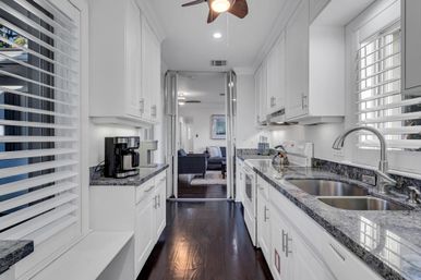 Bright modern galley kitchen with white cabinets, granite countertops, stainless steel double sink, coffee maker, plantation shutters, dark hardwood floors and ceiling fan, opening to a living room.