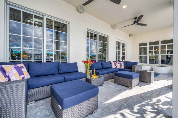 Sunlit covered outdoor patio lounge with navy wicker sectional and ottomans, glass coffee table with flowers and champagne, ceiling fans and large windows reflecting waterfront and string lights.