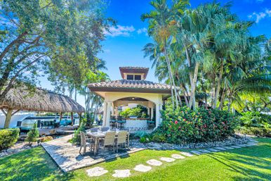 Tropical waterfront backyard with covered patio bar, outdoor dining table on a stone terrace, tiki hut and boat dock framed by palm trees, flowering shrubs and a stone path under a bright blue sky.