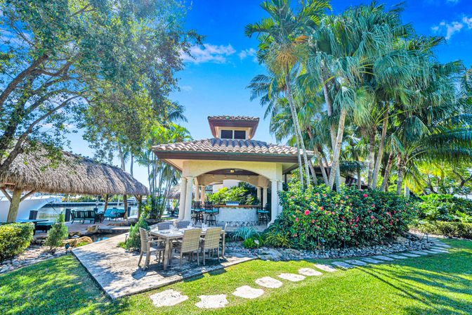 Tropical waterfront backyard with covered patio bar, outdoor dining table on a stone terrace, tiki hut and boat dock framed by palm trees, flowering shrubs and a stone path under a bright blue sky.
