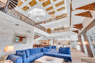 Sunlit grand hotel atrium lounge with plush blue velvet sectional, multi-level balconies and railings, coffered skylight ceiling, spiral staircase and carved wooden decorative brackets