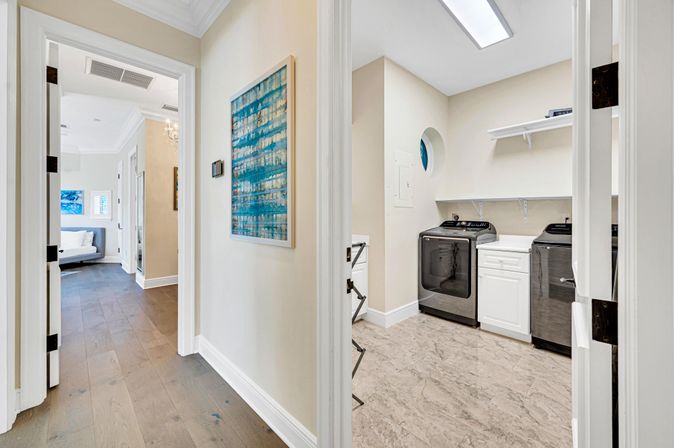 Sunlit hallway opening into a modern laundry room with dark front-loading washer and dryer, white cabinetry and shelves, marble-look tile floor, beige walls and a blue abstract wall painting in the hall.