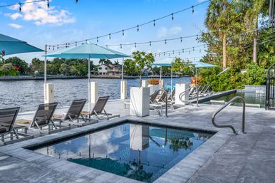 Riverside pool deck with lounge chairs and turquoise umbrellas, string lights overhead, a modern spa and palm trees lining the waterfront