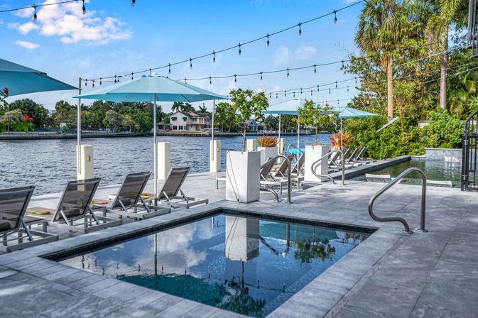 Riverside pool deck with lounge chairs and turquoise umbrellas, string lights overhead, a modern spa and palm trees lining the waterfront