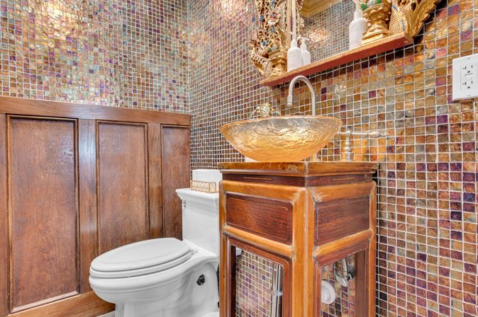 Ornate powder room with iridescent mosaic tile walls, wood wainscoting, white toilet and amber glass vessel sink atop a carved wooden vanity