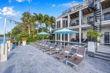 Luxury tropical waterfront villa with poolside lounge chairs and blue umbrellas on a stone terrace, palm trees, second-floor balconies and a spiral staircase overlooking a calm bay