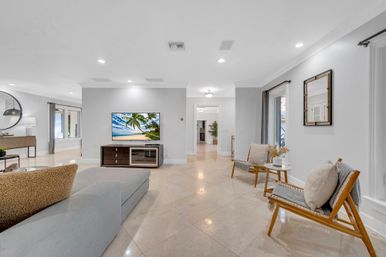 Bright, airy modern open-concept living room with marble tile floors, gray sectional sofa, wooden accent chairs, TV displaying a beach scene, recessed lighting and neutral coastal décor.