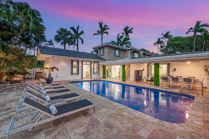 Luxury tropical backyard pool at dusk with blue-tiled rectangular pool, row of chaise lounges with towels, covered outdoor dining and seating, stone-clad two-story house and palm trees under a pink-purple sky