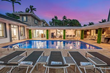 Luxury modern villa backyard at dusk with an illuminated rectangular swimming pool, row of lounge chairs, outdoor dining and seating areas, palm trees and a purple-pink sunset sky.