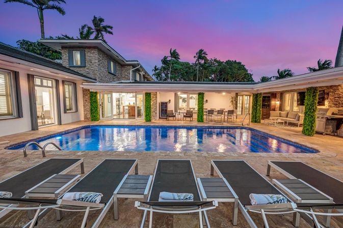 Luxury modern villa backyard at dusk with an illuminated rectangular swimming pool, row of lounge chairs, outdoor dining and seating areas, palm trees and a purple-pink sunset sky.