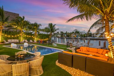 Sunset-lit tropical waterfront patio with pool, palm trees, orange outdoor sofa, dining table and a nearby marina dock