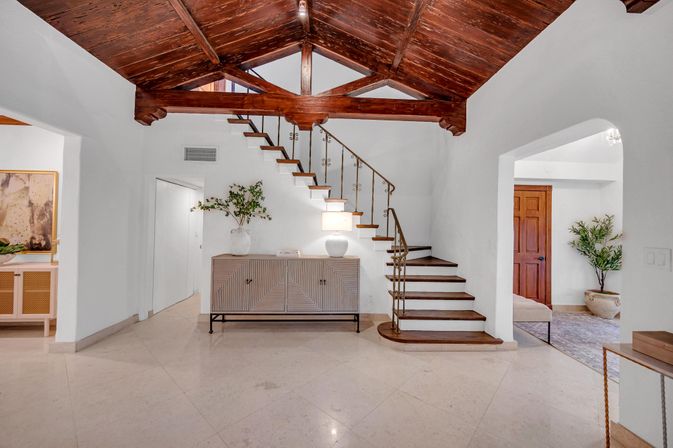Sunlit Mediterranean-style entry with rich wood vaulted ceiling and exposed beams, floating wooden staircase with decorative iron railing, light marble floor, modern console topped with lamp and vase, arched doorways and potted greenery.