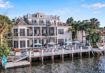 Multi-level luxury waterfront home with wraparound balconies, spiral staircase, palm trees and a private dock lined with lounge chairs and blue umbrellas