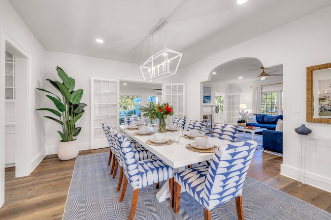 Bright coastal-style dining room with white table, blue patterned upholstered chairs, geometric chandelier, potted tropical plant, hardwood floors, and open view into the living room and backyard.