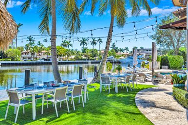 Sunny tropical waterfront patio with al fresco dining tables and chairs on green lawn under palm trees and string lights, docked yacht on a calm canal.