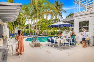 Sunny tropical backyard pool patio with palm trees, navy umbrella, outdoor dining set, lounge chairs and guests enjoying drinks.
