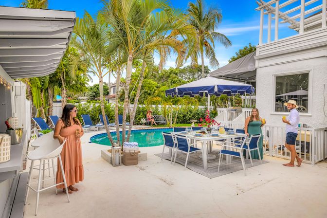 Sunny tropical backyard pool patio with palm trees, navy umbrella, outdoor dining set, lounge chairs and guests enjoying drinks.